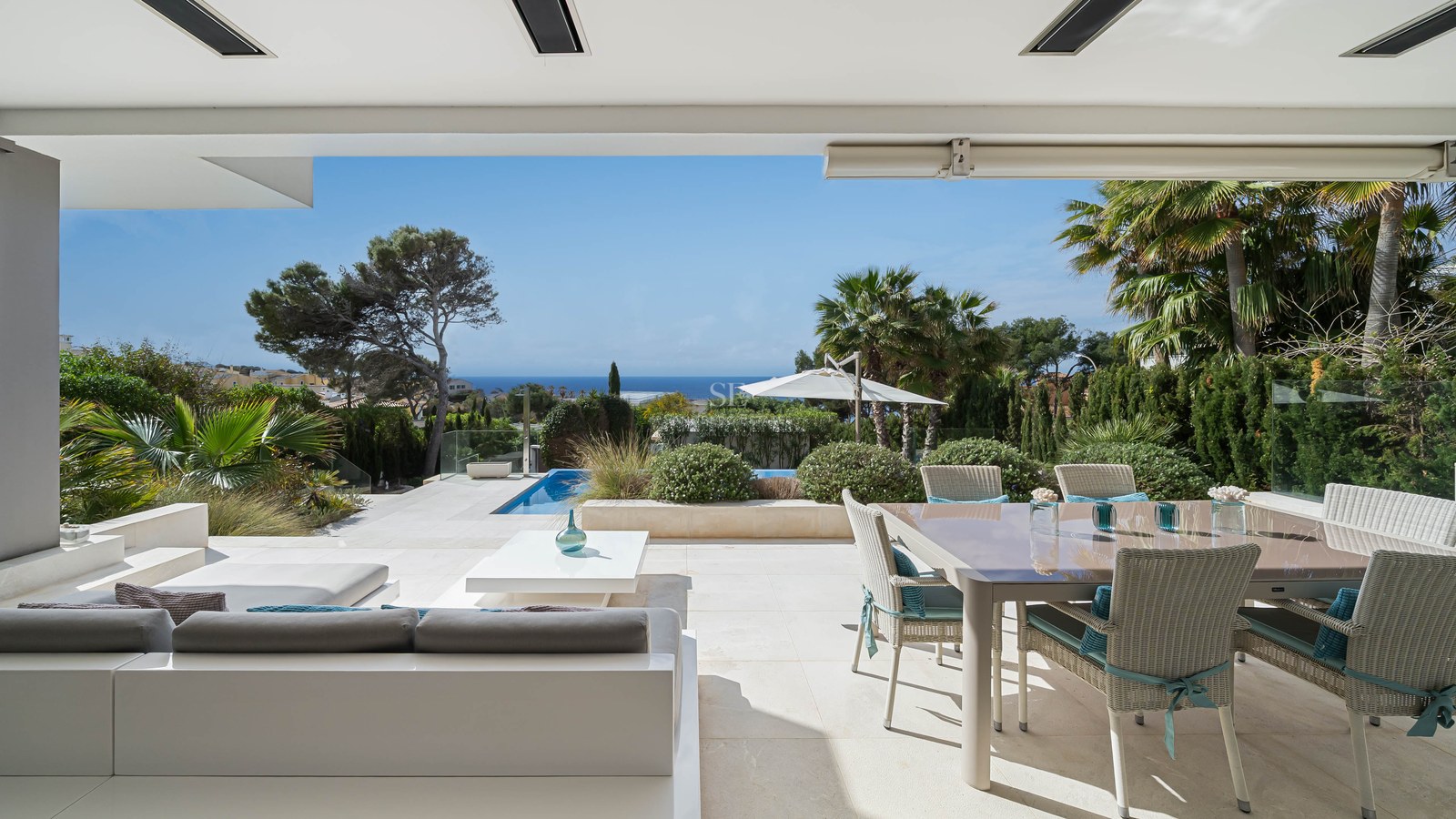 Spacious luxury terrace with white lounge seating, wicker dining set, stone floors, and a view of the pool and sea.