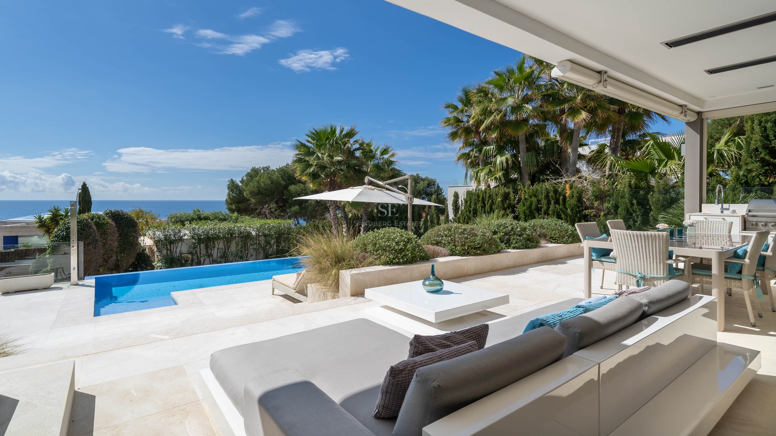 Luxury terrace with white lounge furniture, stone flooring, infinity pool, and palm trees overlooking the sea.