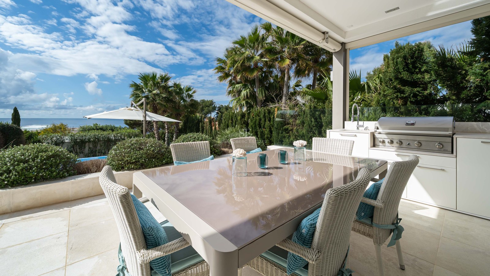 Outdoor dining table and integrated BBQ kitchen on a stone terrace overlooking the sea and palm trees.