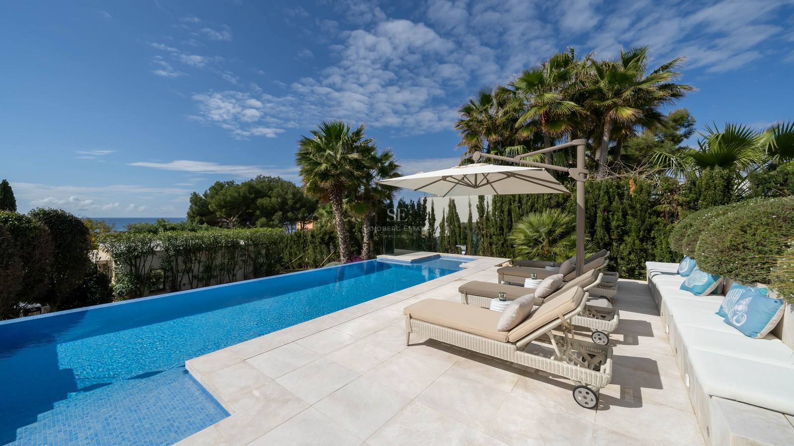 A sun-drenched terrace featuring an infinity pool, beige sun loungers under an umbrella, and distant sea views.