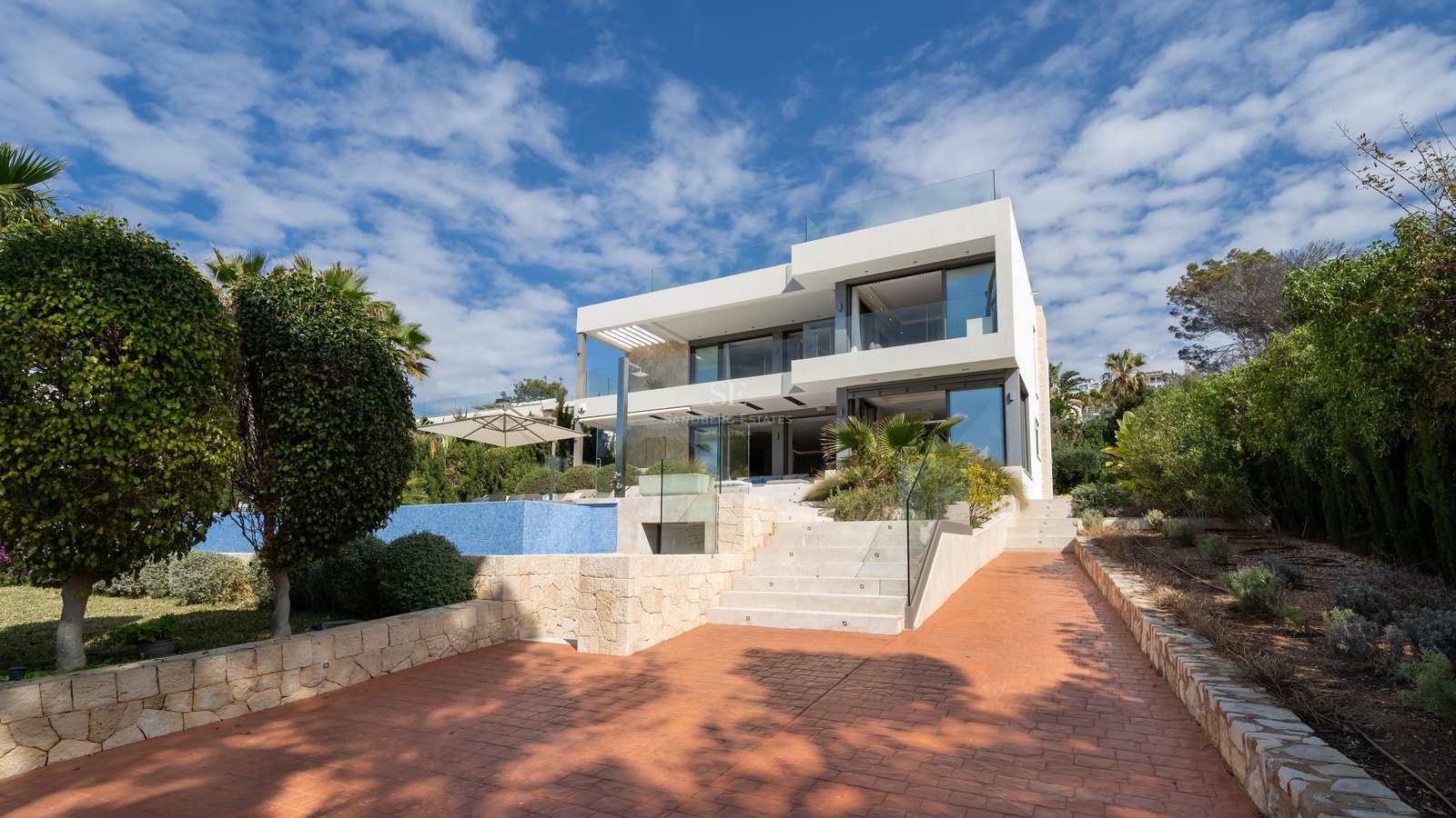 Three-story modern white villa with glass balconies, blue swimming pool, and stone stairs under a bright blue sky.