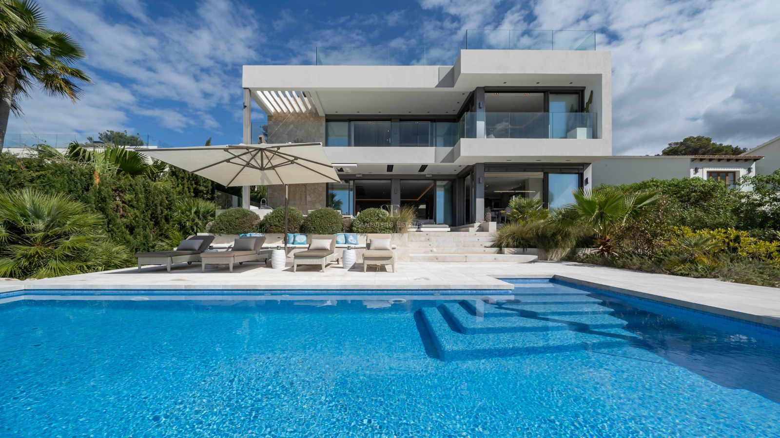 Modern two-story white villa with large swimming pool, sun loungers, and glass balconies under a blue sky.