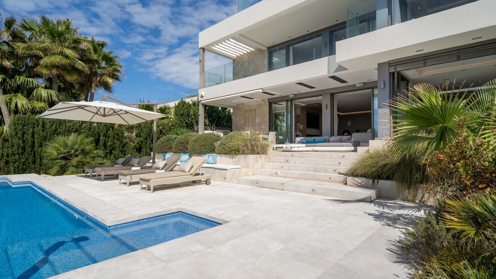 Modern villa featuring a blue tiled swimming pool, designer sun loungers, and natural stone facade under a clear sky.