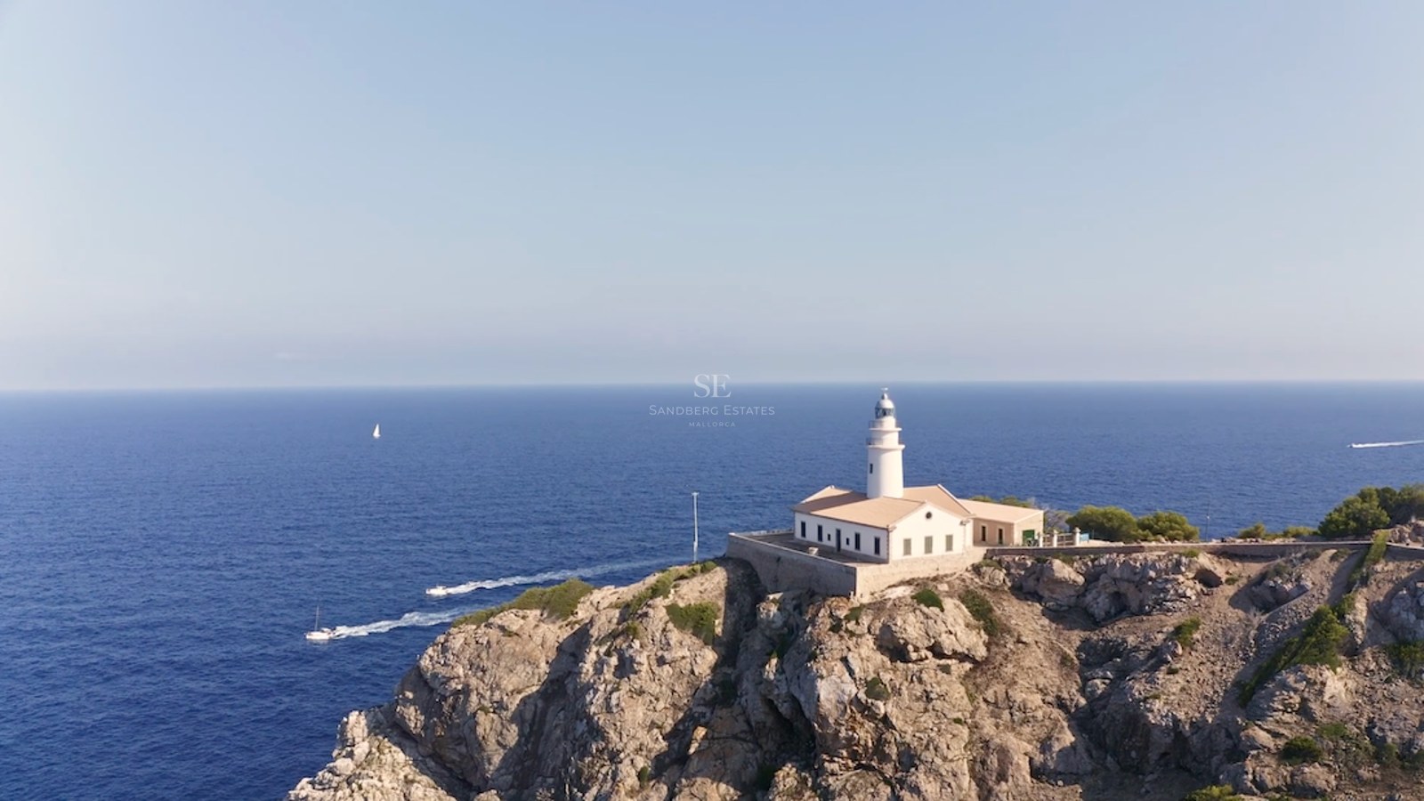 Ein weißer Leuchtturm auf einer zerklüfteten Felsklippe mit Blick auf das tiefblaue Mittelmeer und Boote.