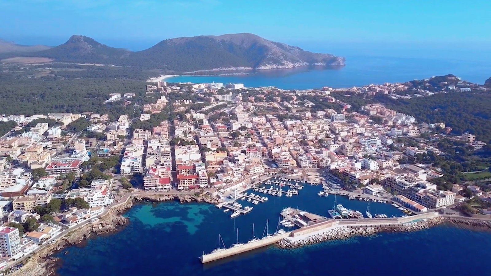 Luftaufnahme eines mediterranen Yachthafens mit Booten, türkisblauem Wasser, einer Küstenstadt und Bergen im Hintergrund.