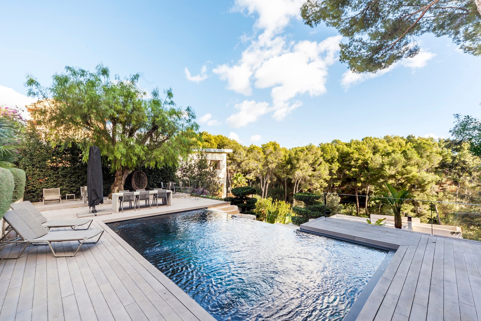 Piscine à débordement de luxe entourée d'une terrasse en bois et d'arbres verdoyants sous un ciel bleu clair.