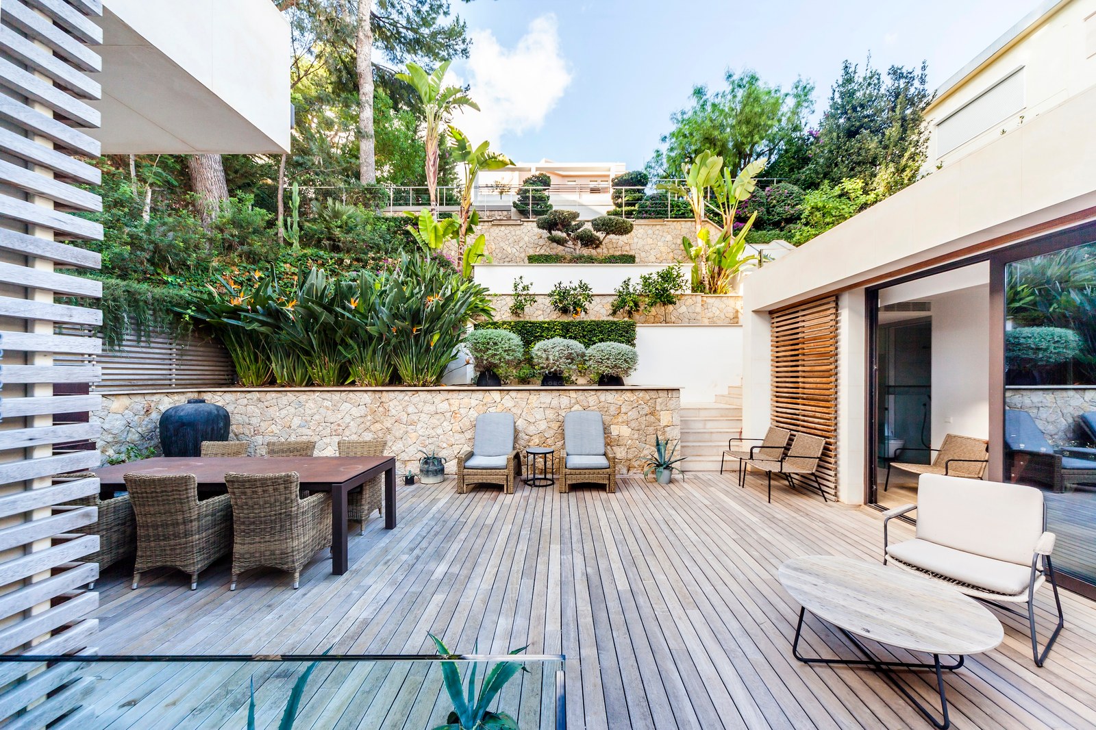 Grande terrasse en bois avec mobilier en osier devant un mur de pierre à plusieurs niveaux et plantes tropicales.