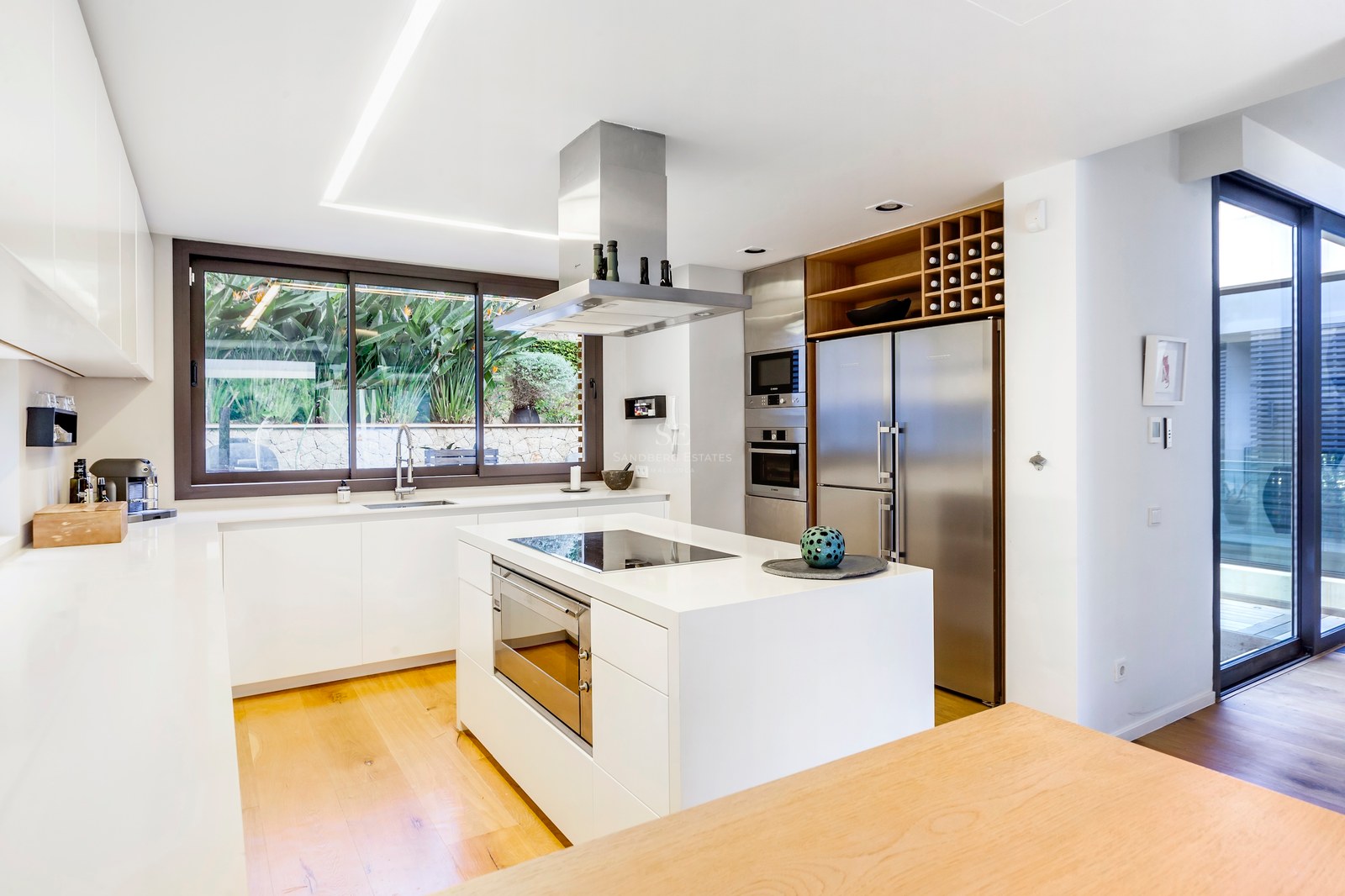 Cuisine blanche moderne avec îlot central, appareils en acier inoxydable et parquet en bois avec vue sur le jardin.