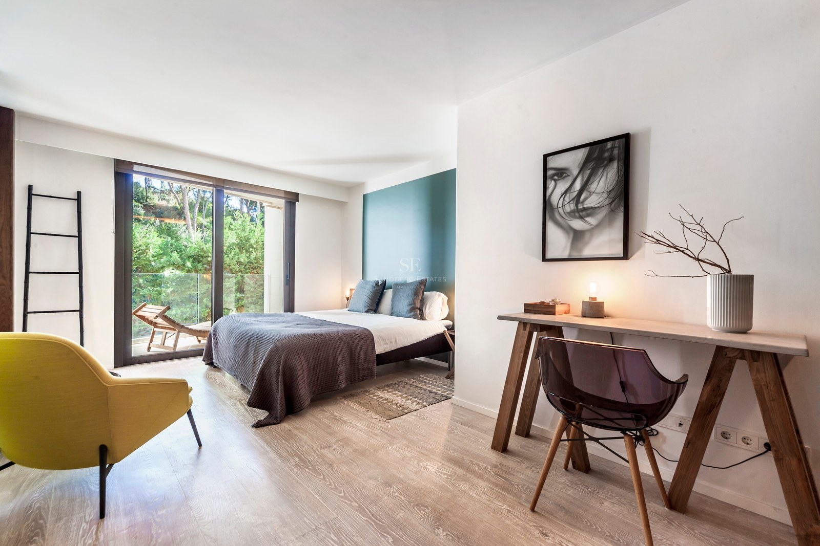 Chambre moderne avec mur sarcelle, bureau en bois, fauteuil jaune et baies vitrées ouvrant sur une terrasse verdoyante.