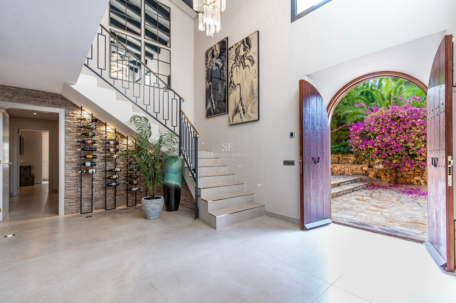 Vestíbulo de lujo con escalera moderna, botellero integrado y puertas de arco abiertas hacia un patio con buganvillas.