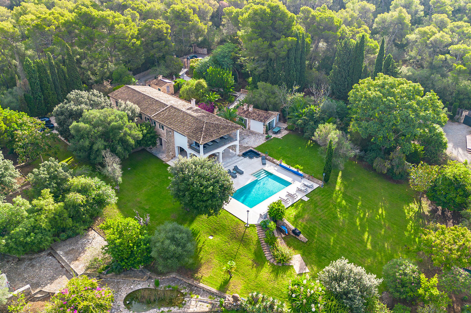 Vista aérea de una gran villa de piedra rodeada de bosque, con piscina turquesa y amplios jardines verdes.