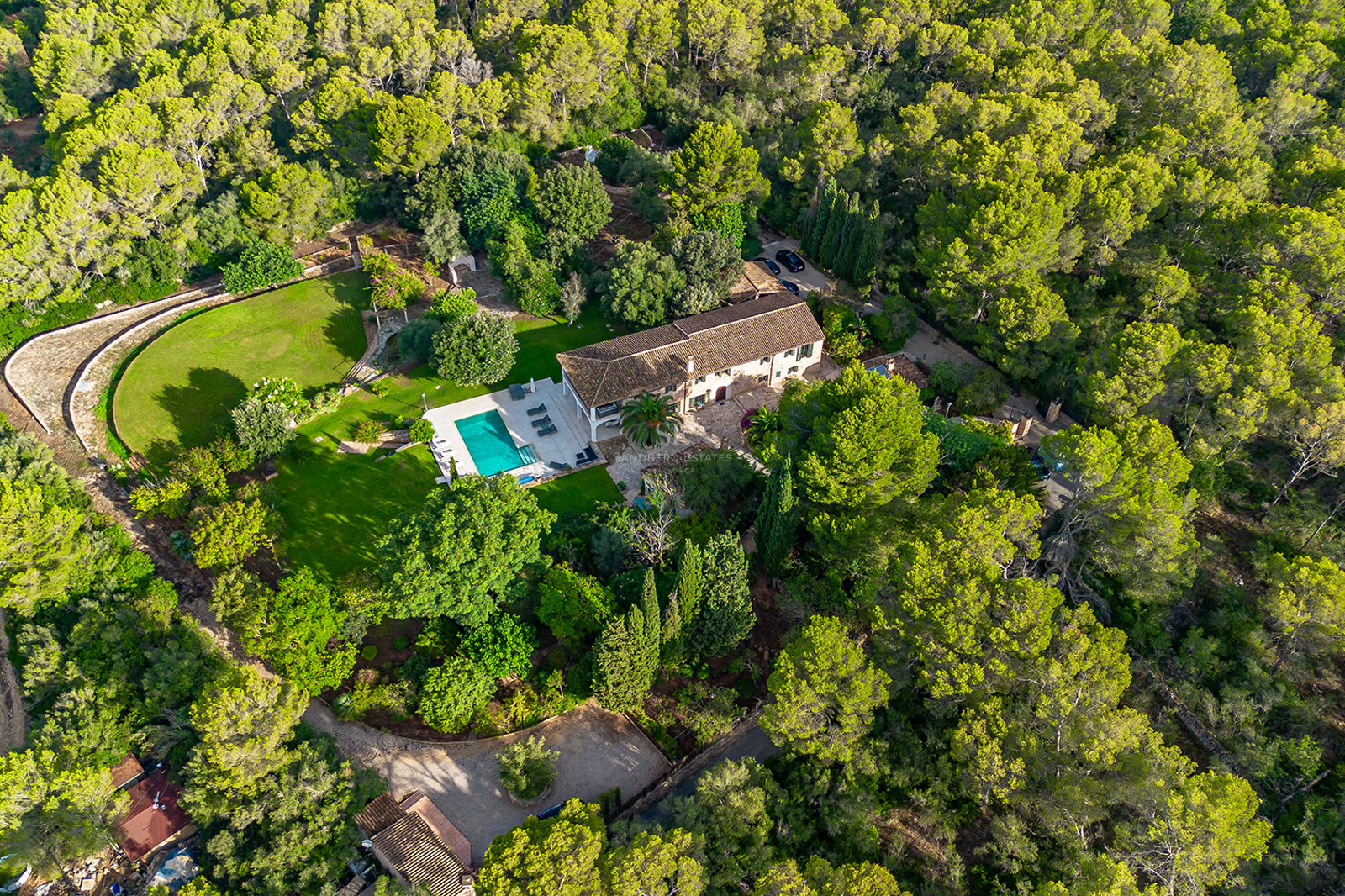 Vista aérea de una villa tradicional con piscina turquesa rodeada por un denso bosque de pinos y jardines extensos.
