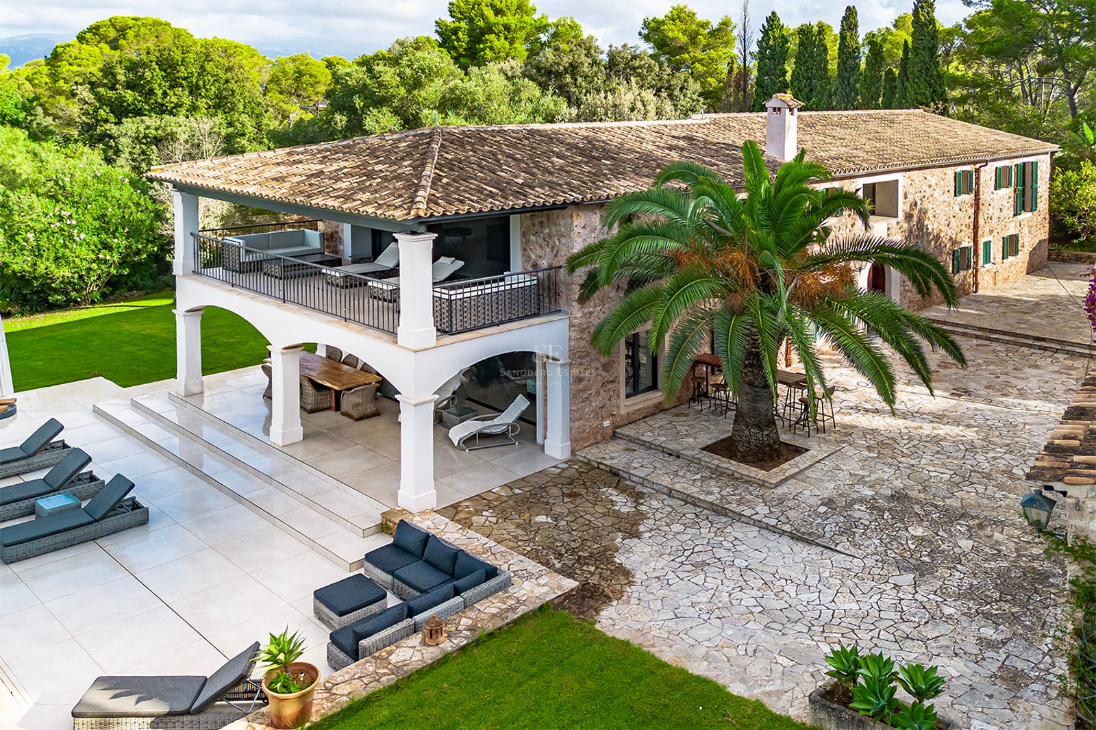 Vista aérea de una villa de piedra con arcos blancos, terrazas exteriores, una palmera y zona de piscina.