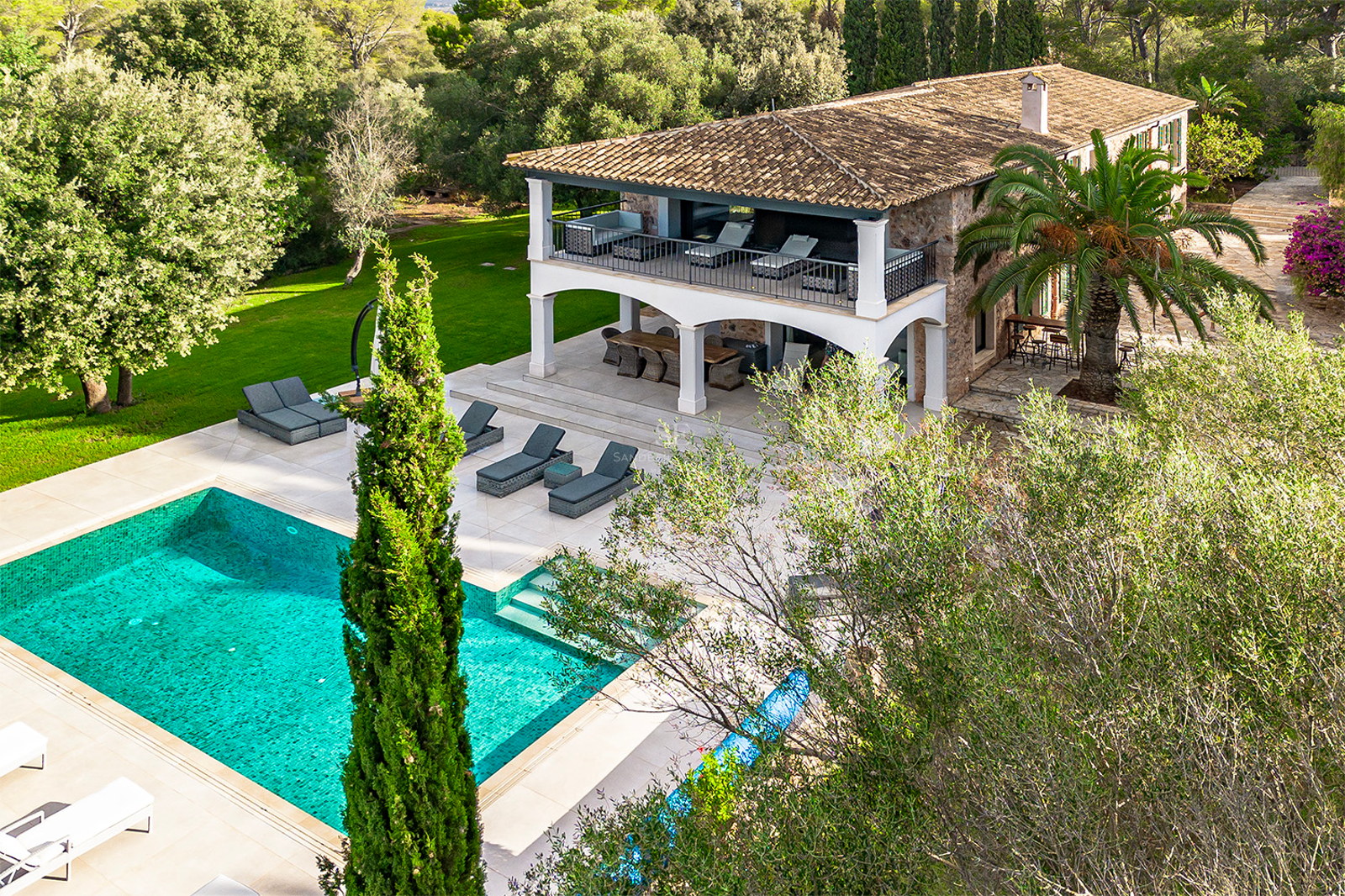 Vista elevada de una villa de piedra con piscina turquesa, tumbonas y jardines mediterráneos frondosos.