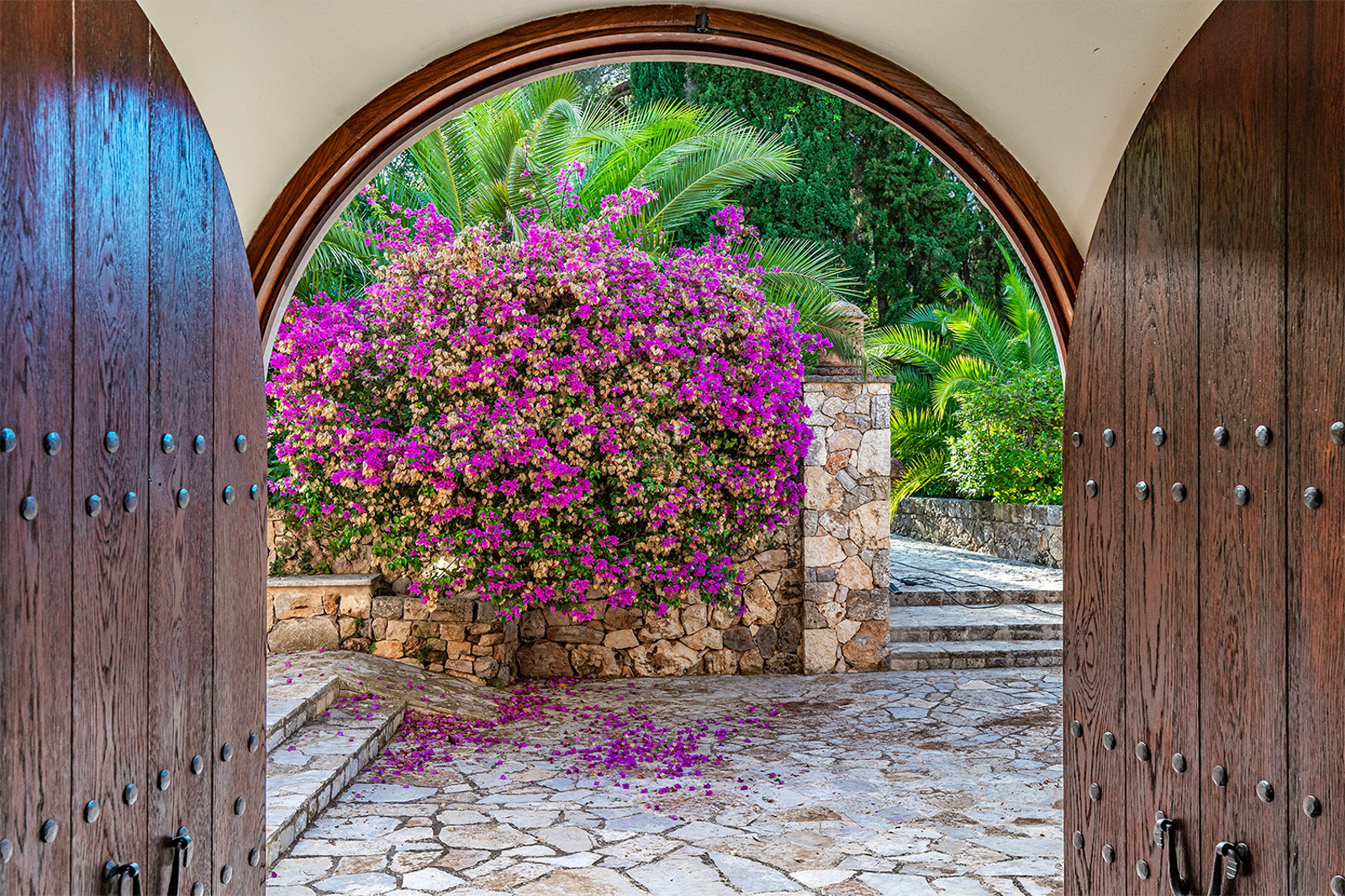 Vista desde puertas de madera tallada hacia un patio de piedra con buganvillas púrpuras y vegetación exuberante.