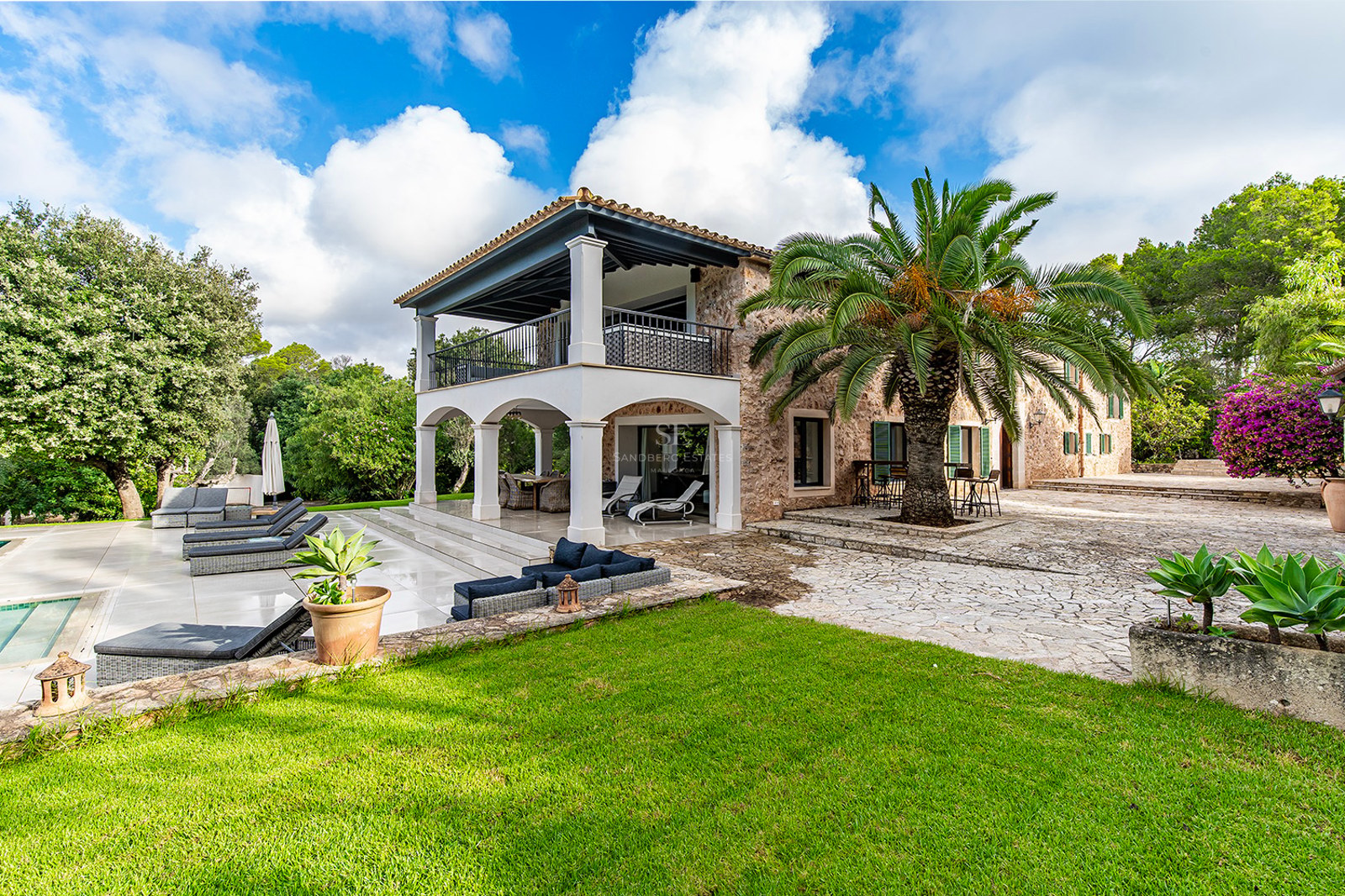 Villa de piedra natural de dos niveles con arcos, palmera central, jardín cuidado y zona de piscina.
