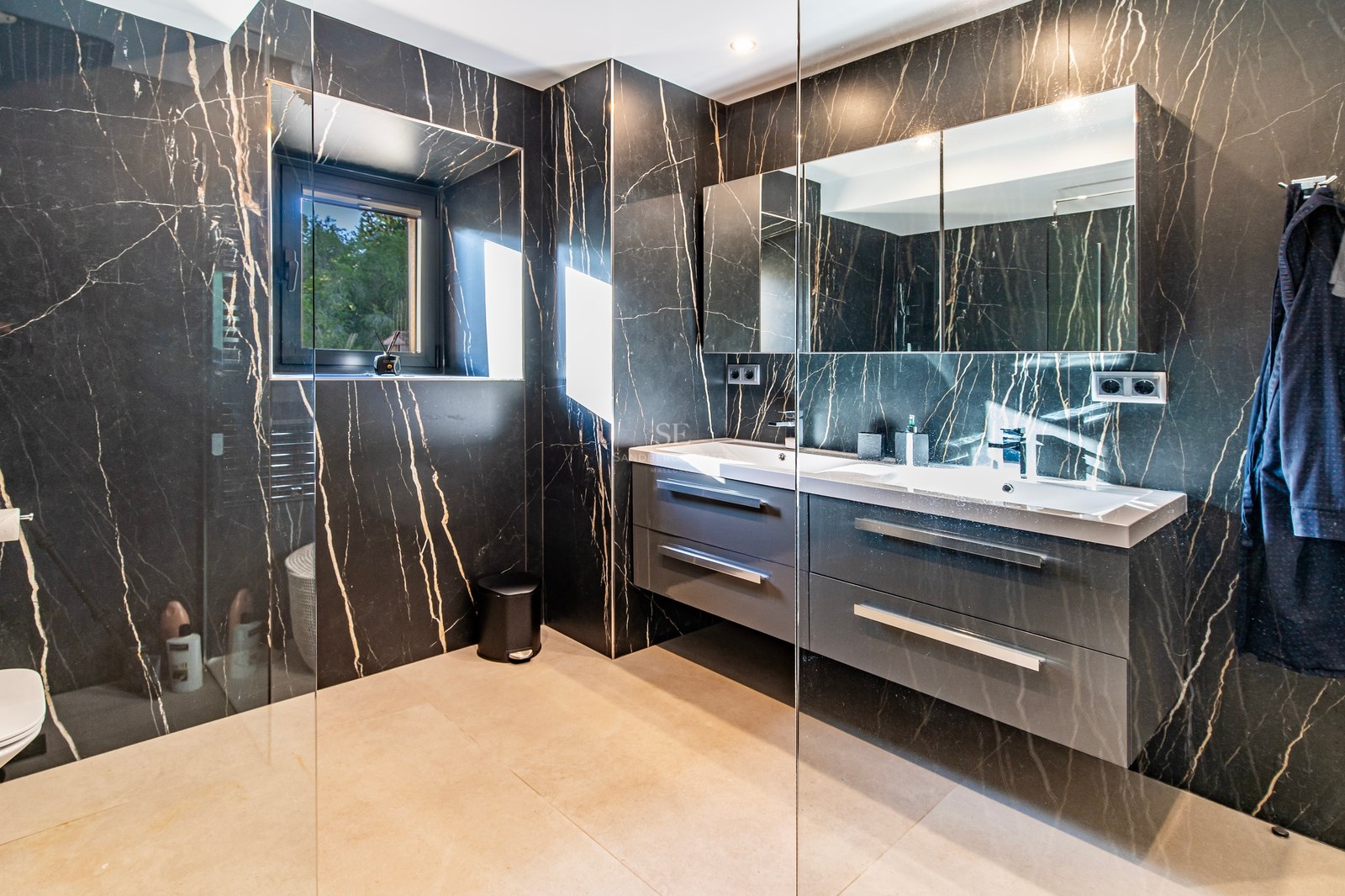 Baño moderno con paredes de mármol negro veteado, mueble de doble lavabo gris y ducha con mampara de cristal.
