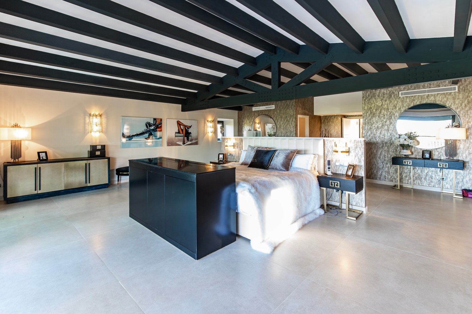 Amplio dormitorio principal con vigas de madera negras, cama king con manta de piel y suelos de piedra clara.