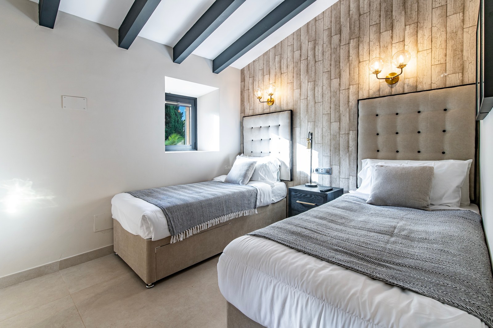 Dormitorio contemporáneo con dos camas individuales, cabeceros tapizados, pared de madera y vigas oscuras en el techo.