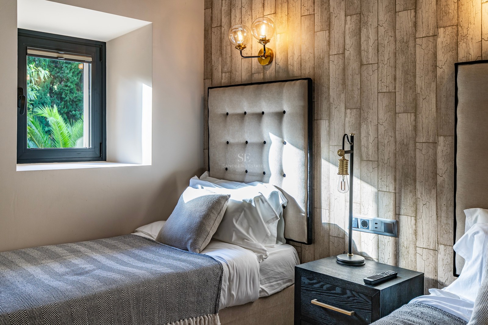 Dormitorio moderno con dos camas, cabecero de tela acolchado, pared con textura de madera y lámparas industriales.