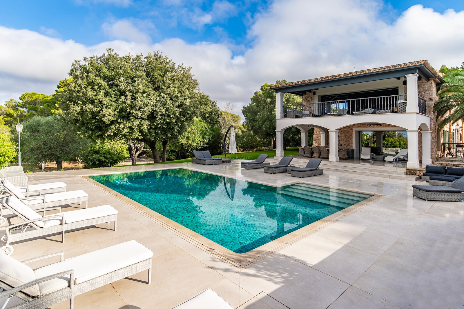 Piscina y terraza de una villa de lujo con fachada de piedra y jardín frondoso bajo un cielo azul.