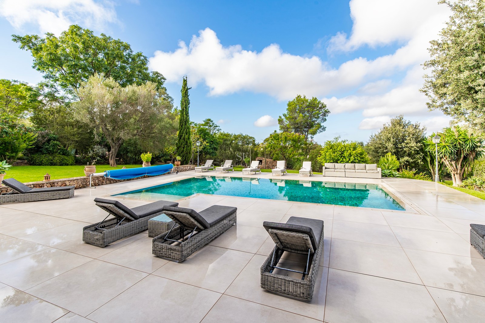 Piscina rectangular azul con amplia terraza de baldosas beige, tumbonas de ratán y jardín frondoso con césped.