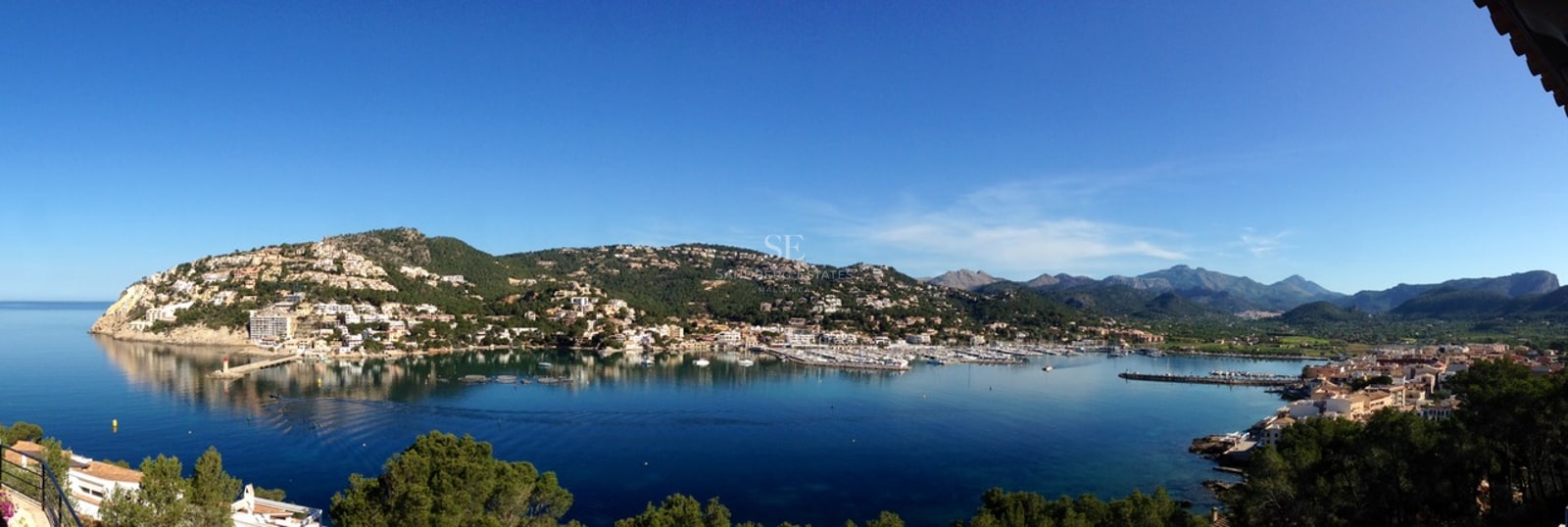 Vue panoramique d'un port méditerranéen avec des yachts, entouré de collines et de montagnes au loin.