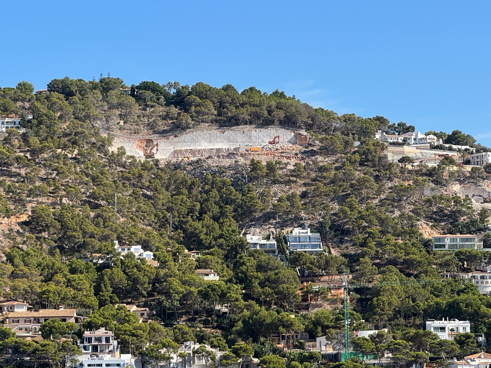 Vue d'une colline verdoyante avec des villas blanches modernes et un chantier de construction sous un ciel bleu.
