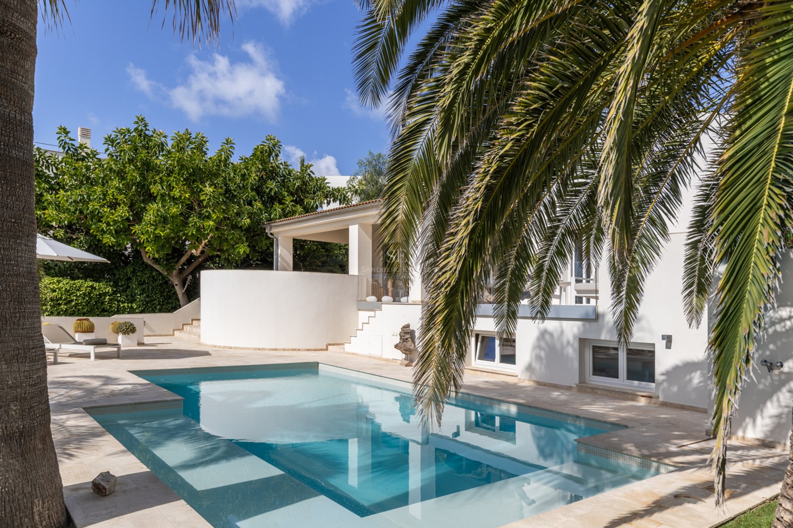 Villa blanche moderne avec piscine rectangulaire entourée de dalles en pierre et de palmiers sous un ciel bleu.