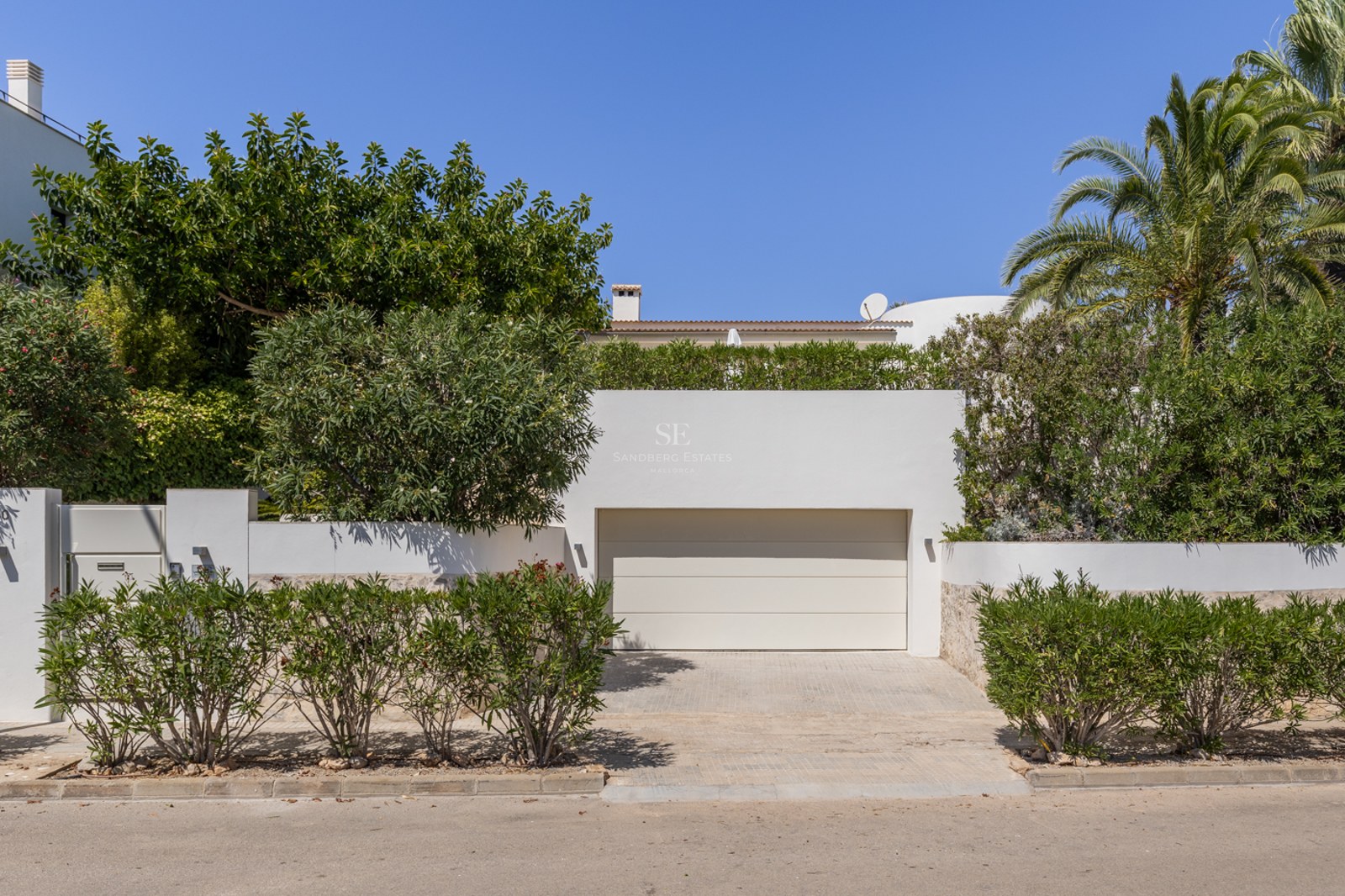 Porte de garage blanche moderne avec allée pavée, entourée de haies vertes et de palmiers sous un ciel bleu.