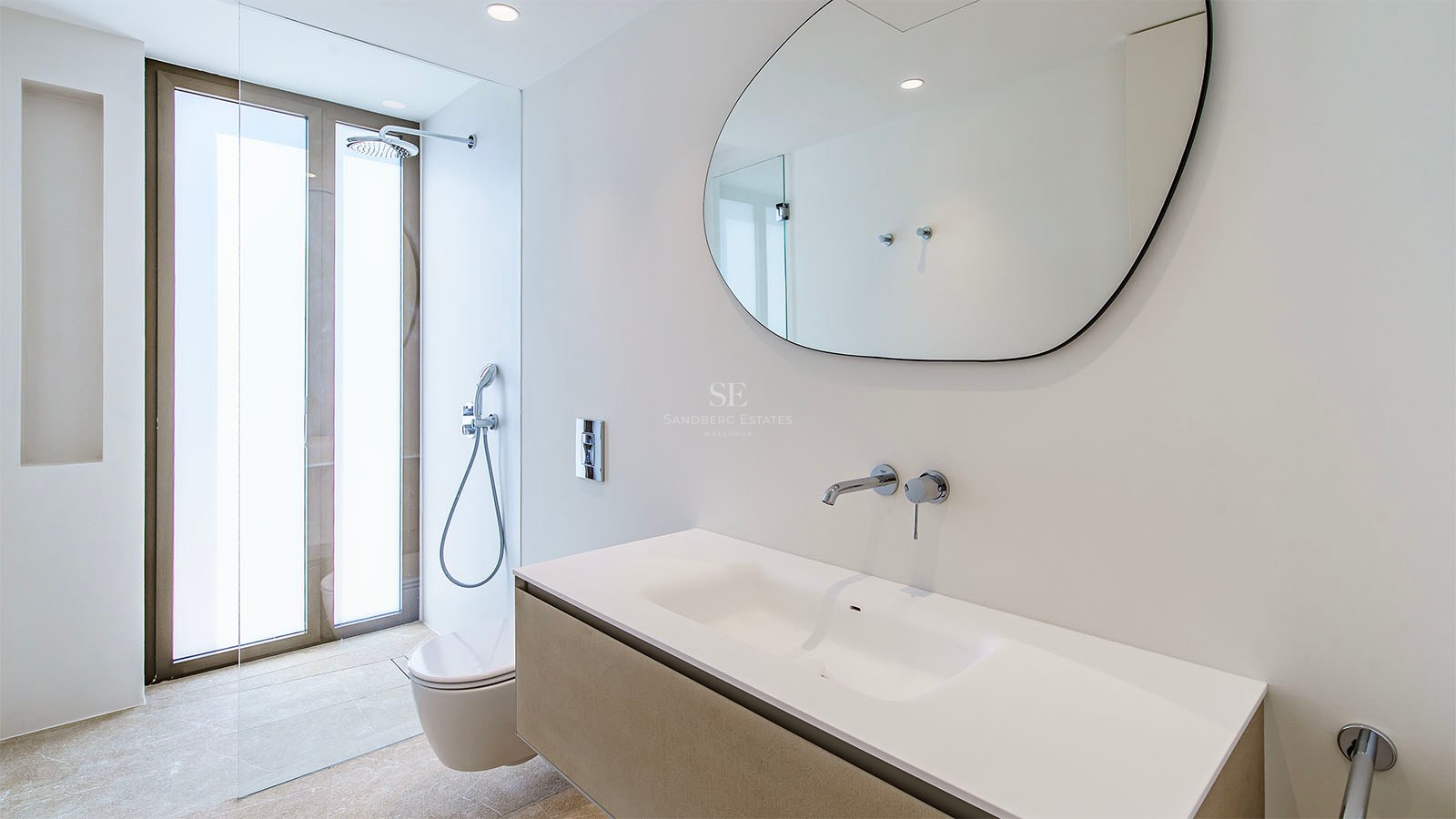 Salle de bain moderne dotée d'un miroir de forme organique, d'un meuble vasque blanc et de robinets chromés encastrés.