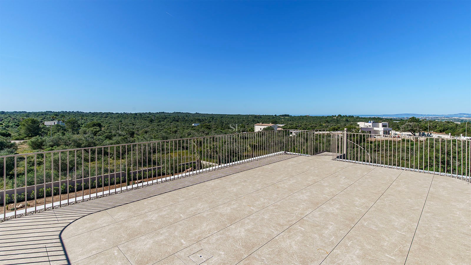 Terrasse en pierre avec garde-corps métallique surplombant un paysage verdoyant sous un ciel bleu azur.