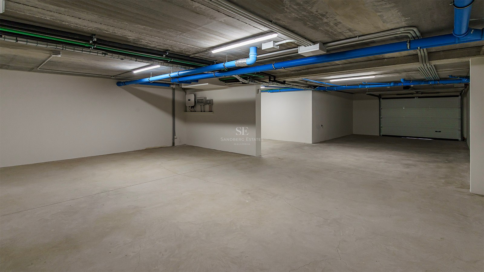 Garage souterrain vide avec sols en béton, tuyaux bleus et murs blancs sous éclairage LED.