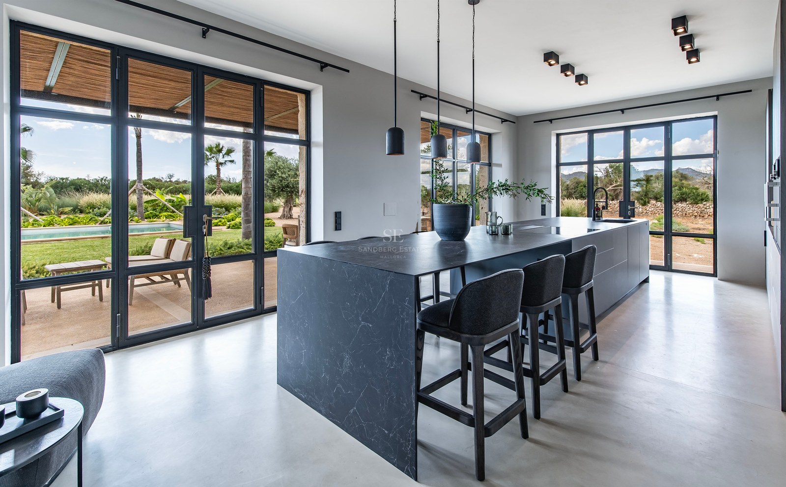 Cuisine moderne avec îlot en marbre noir, tabourets noirs et baies vitrées donnant sur un jardin et une piscine.