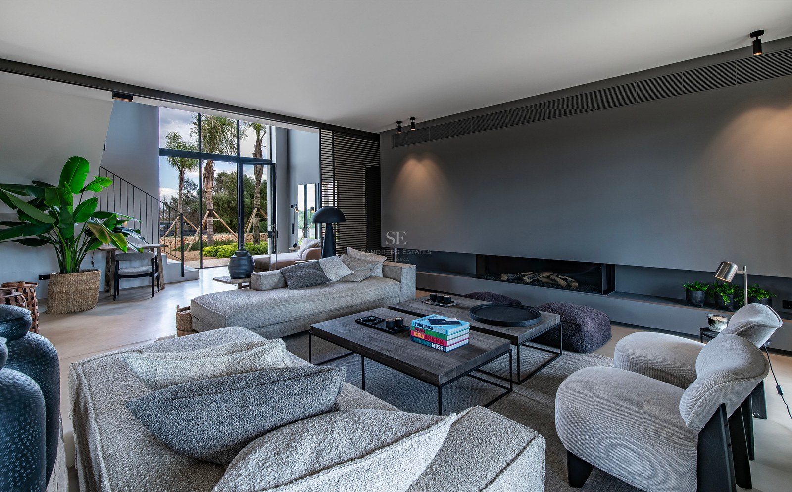 Grand salon moderne avec canapés gris, cheminée intégrée et grandes baies vitrées donnant sur des palmiers.