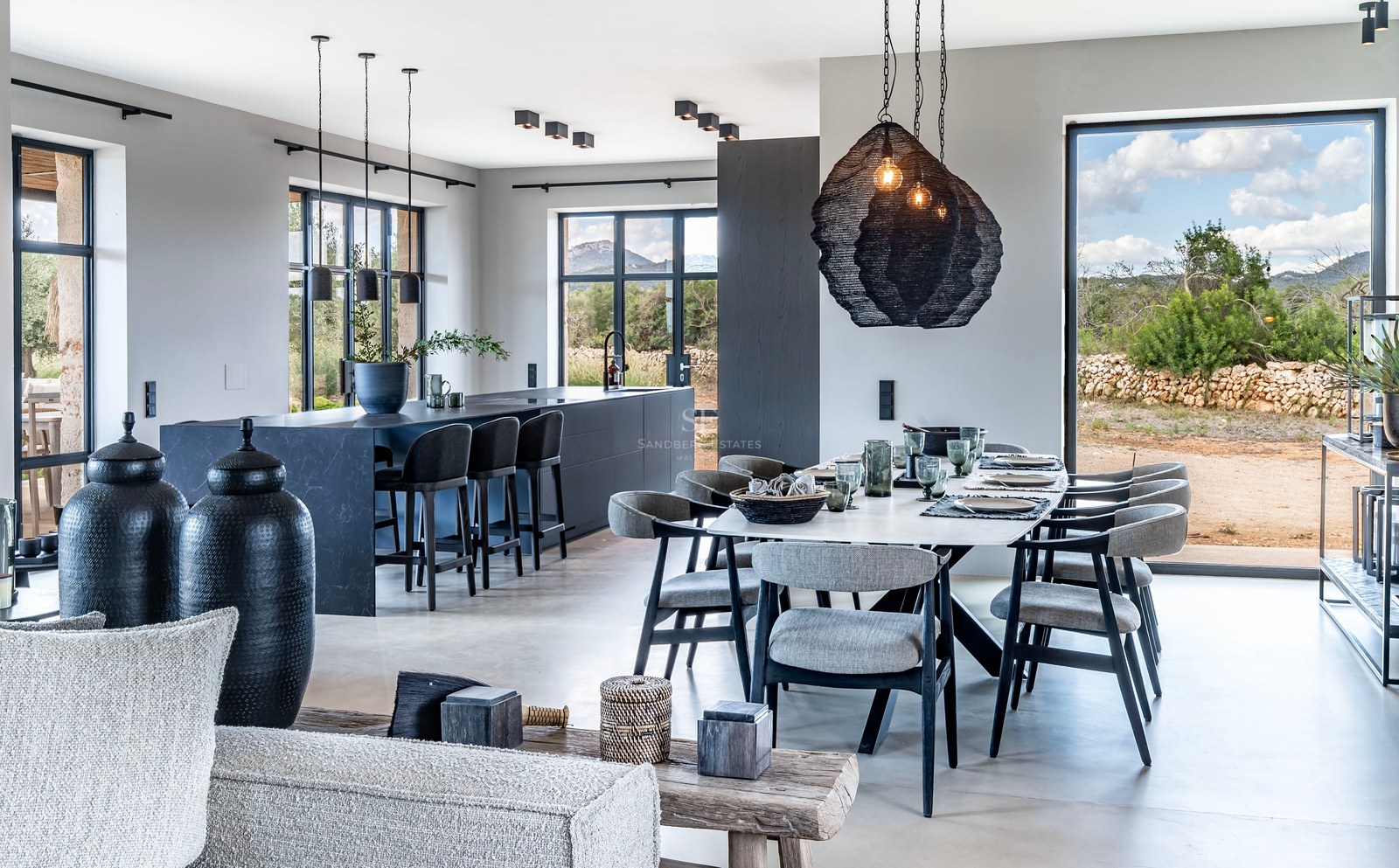 Salle à manger élégante avec une longue table, un îlot de cuisine sombre et de grandes fenêtres donnant sur la campagne.