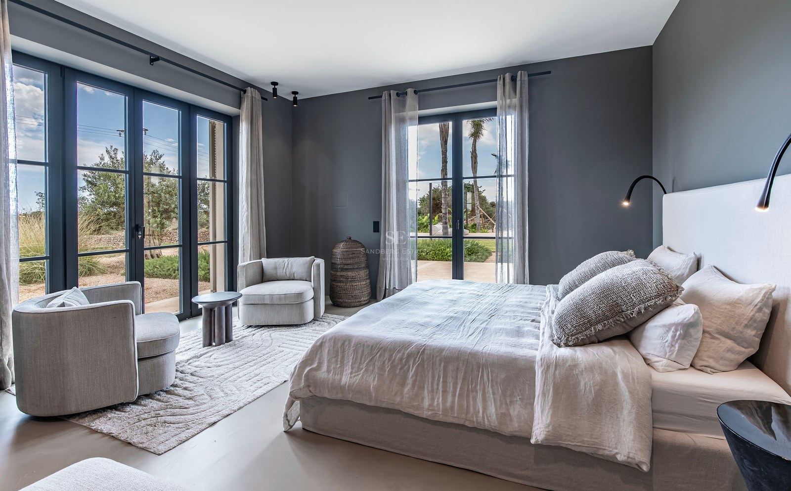 Chambre élégante aux murs gris, grand lit, fauteuils et baies vitrées ouvrant sur l'extérieur.