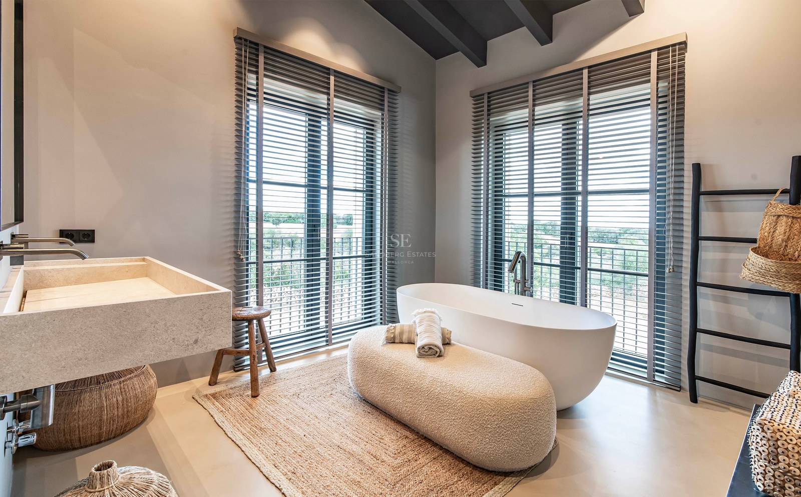 Salle de bain moderne avec baignoire îlot blanche, lavabo en pierre et grandes fenêtres à stores gris.