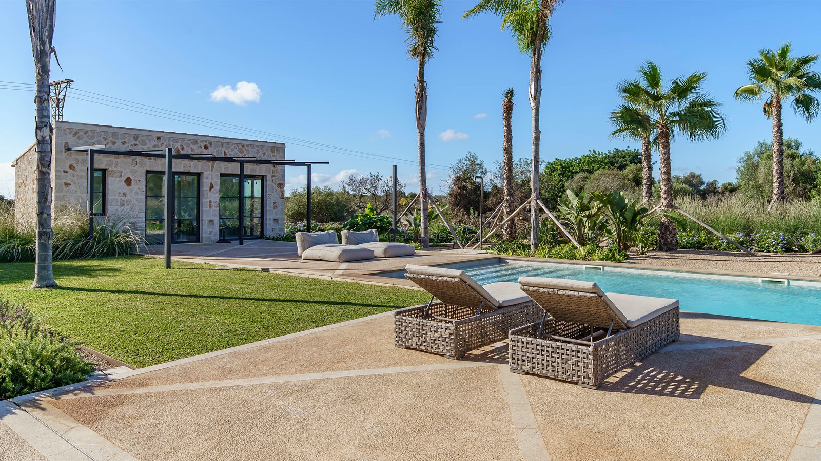 Piscine de luxe avec bains de soleil, palmiers et pavillon en pierre naturelle sous un ciel dégagé.