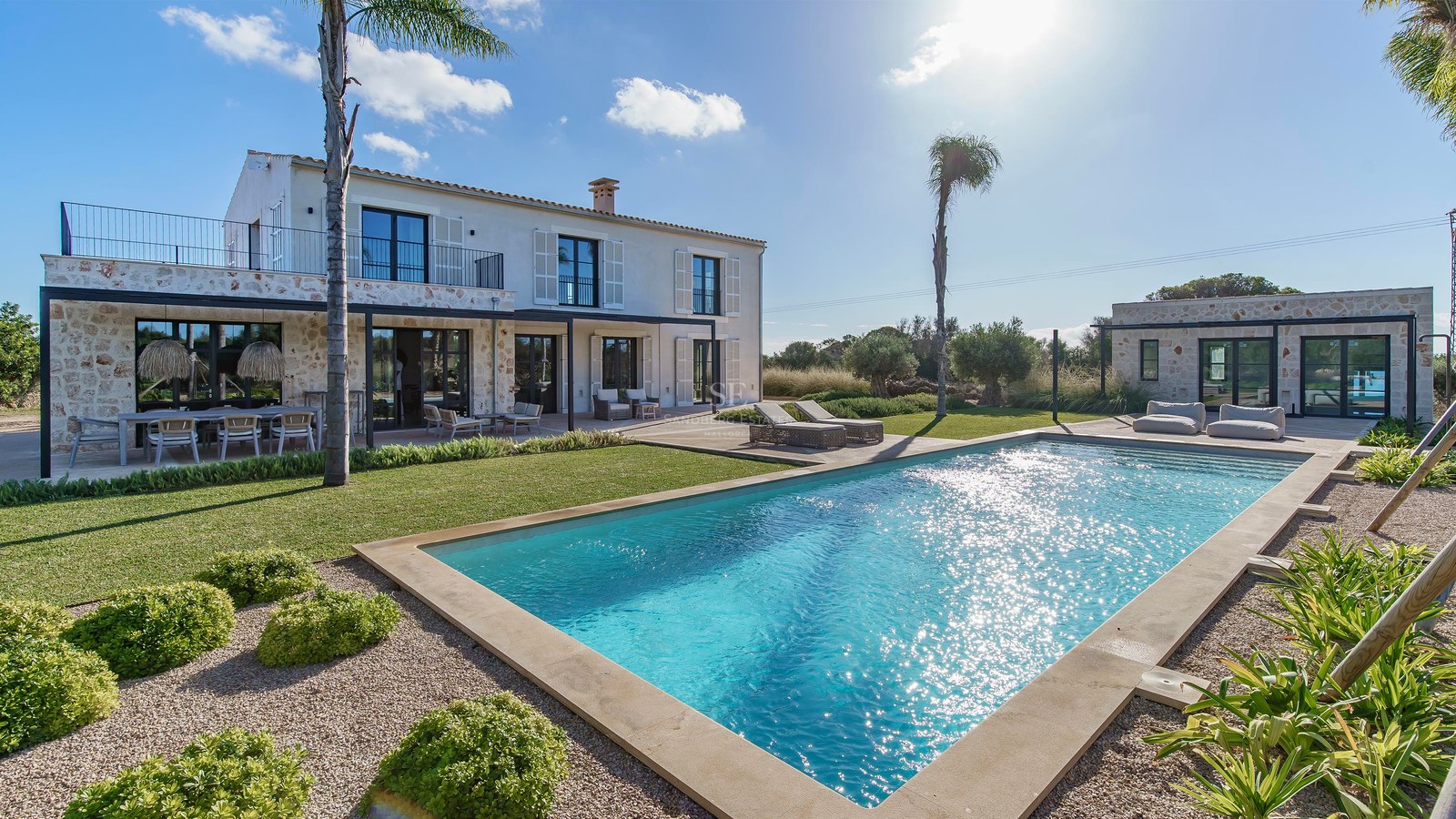 Villa de luxe en pierre avec grande piscine rectangulaire, pelouse impeccable et palmiers sous un soleil éclatant.