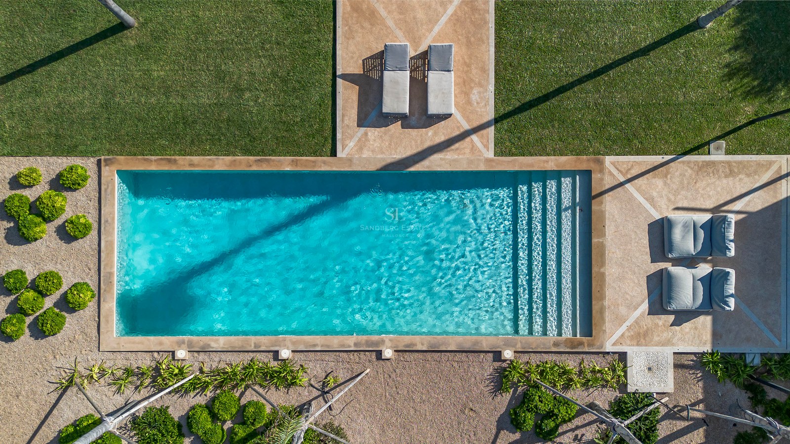 Vue aérienne d'une piscine rectangulaire turquoise entourée d'une pelouse verte, de terrasses en pierre et de transats.