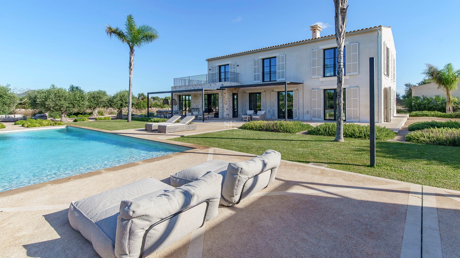 Grande piscine turquoise devant une villa en pierre avec palmiers et pelouse sous un ciel bleu azur.