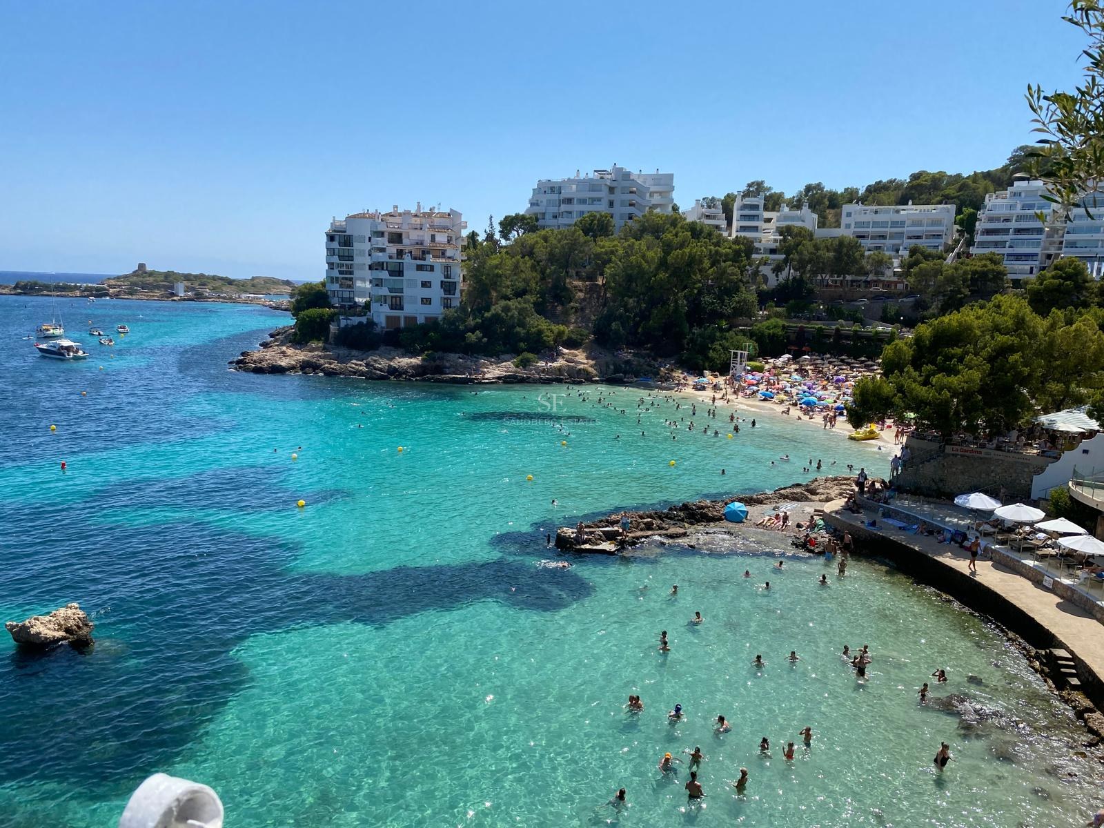 Vista elevada de una bahía turquesa con bañistas, edificios blancos en la costa y acantilados con pinos.
