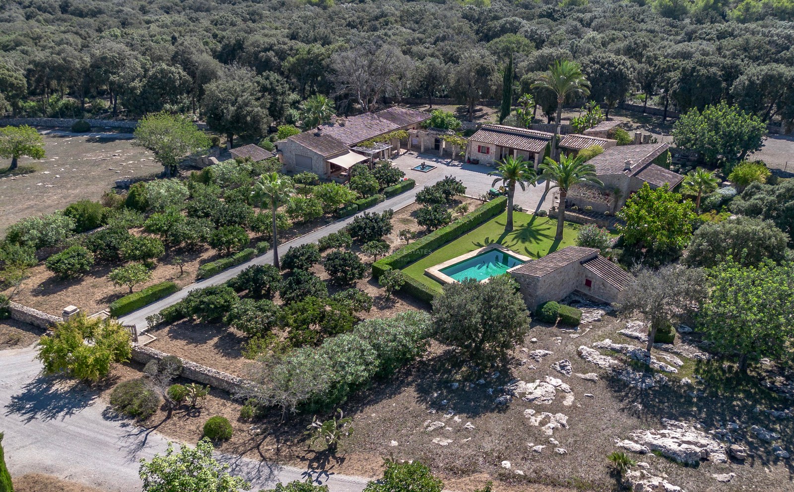 Vue plongeante d'un domaine en pierre avec piscine, palmiers et jardins luxuriants entourés de forêt.