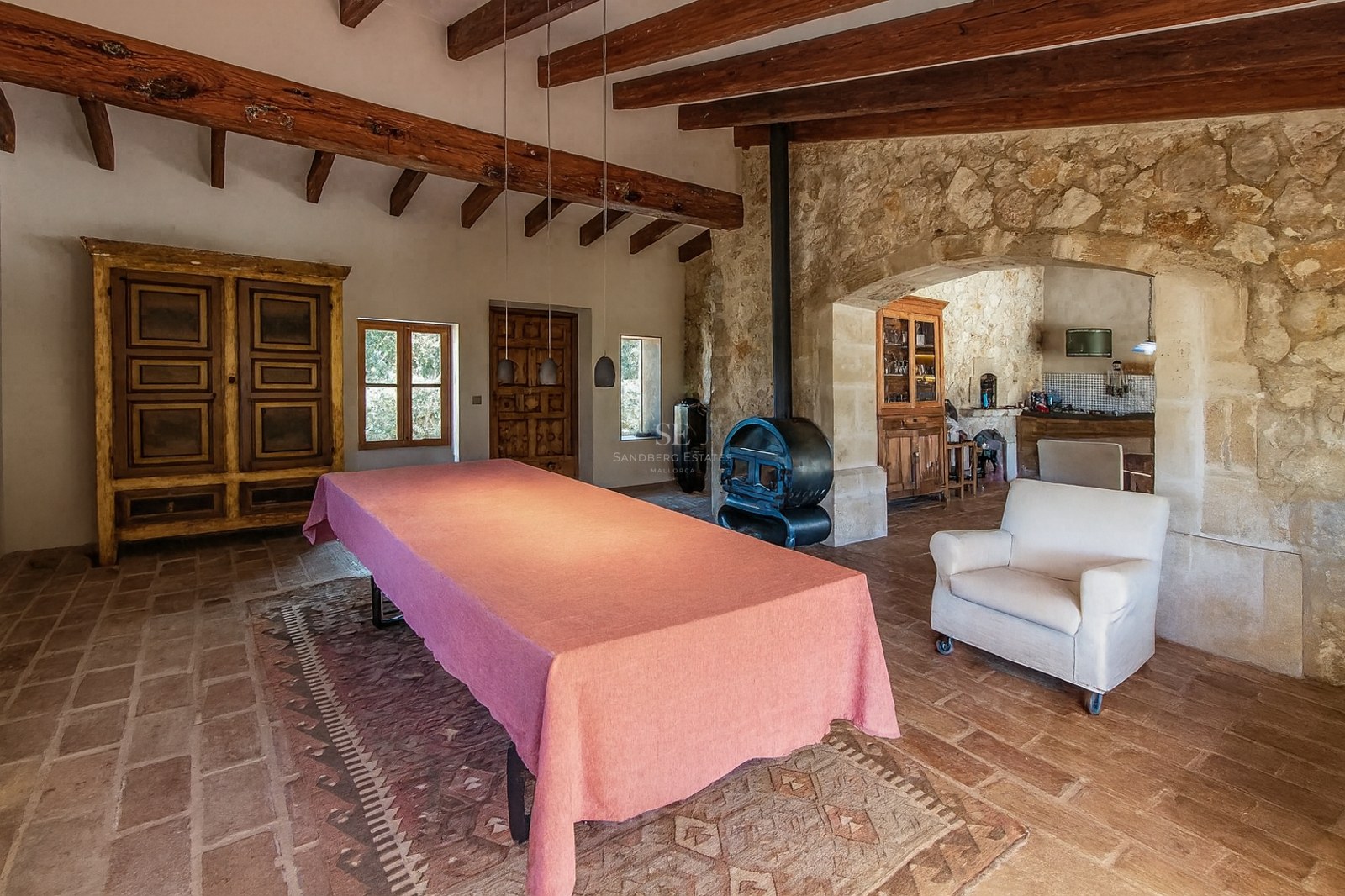 Grande table nappée de rose dans une pièce rustique avec poutres apparentes, murs en pierre et poêle à bois.
