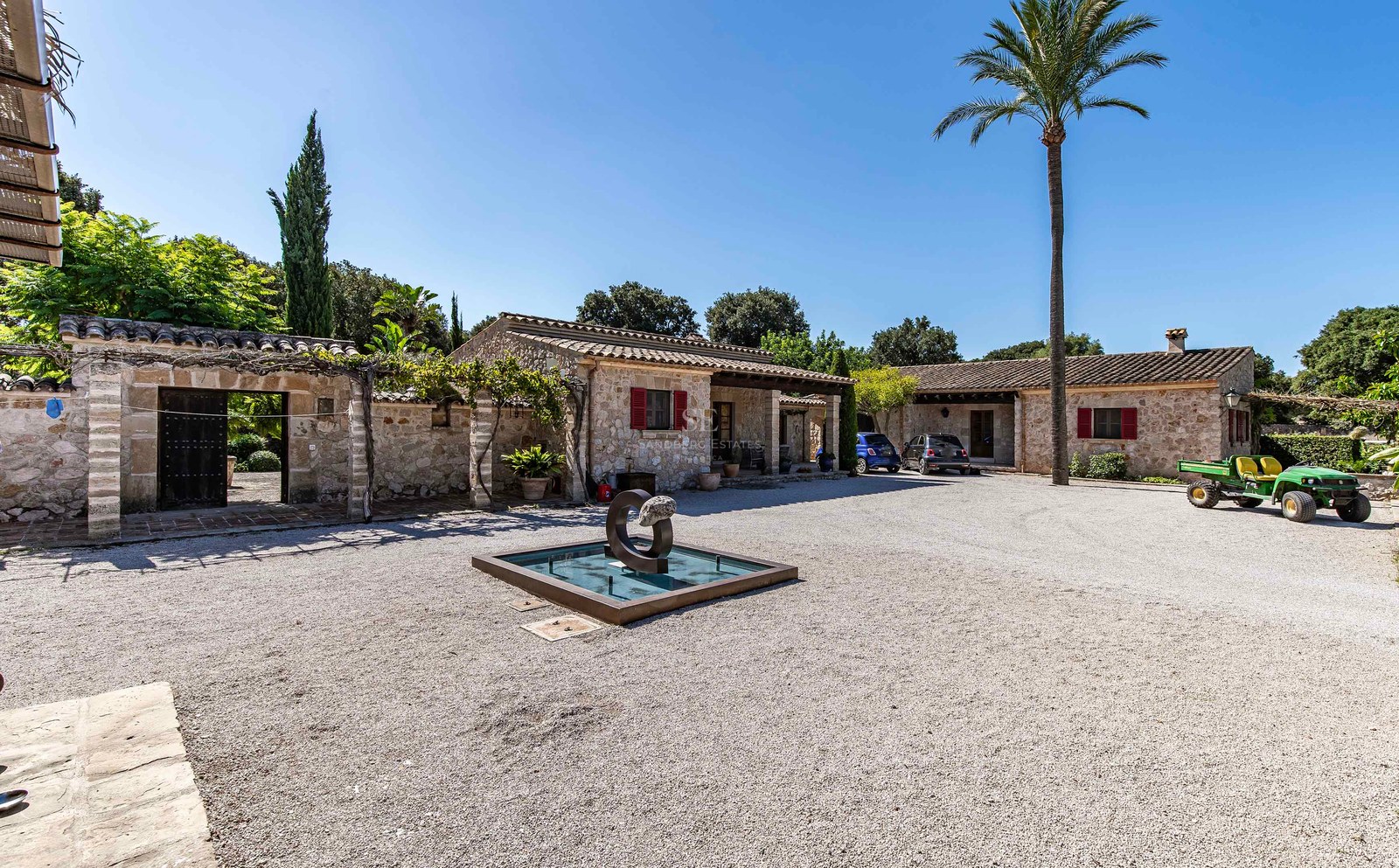 Villa traditionnelle en pierre avec volets rouges, cour en gravier et fontaine moderne sous un ciel bleu.