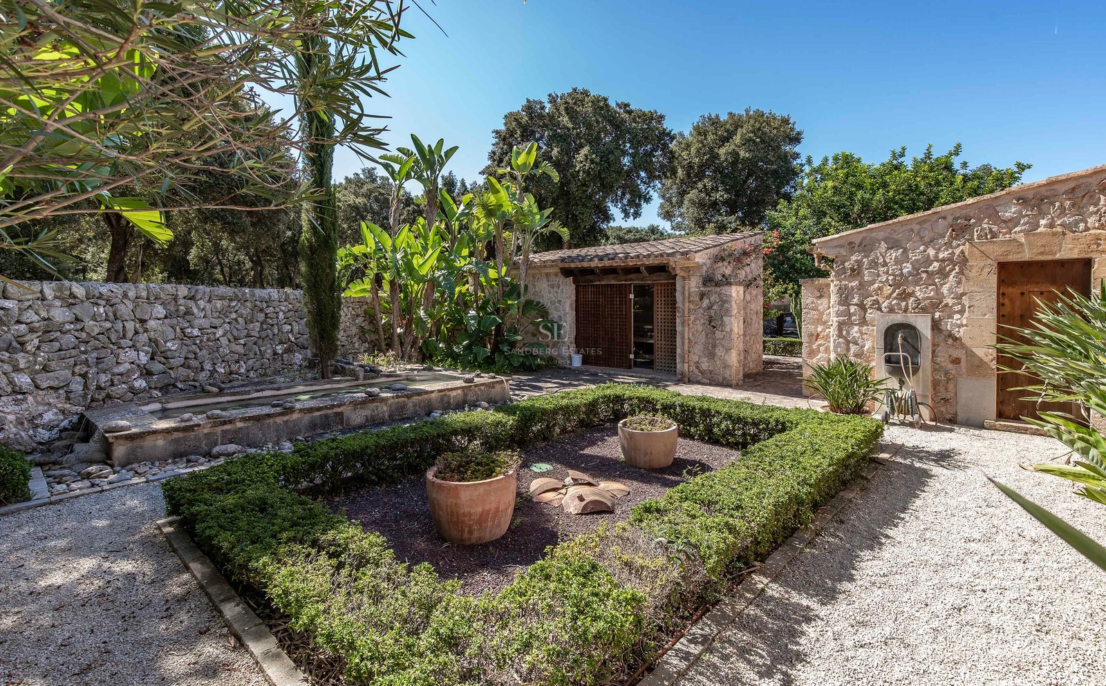 Jardin méditerranéen avec murs en pierre, allées de gravier et dépendances sous un ciel bleu clair.