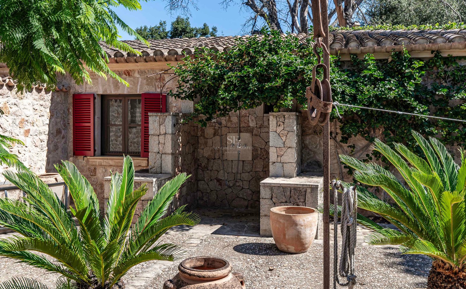 Espace de douche extérieure en pierre avec volets rouges, pots en terre cuite et plantes vertes dans une cour méditerranéenne.
