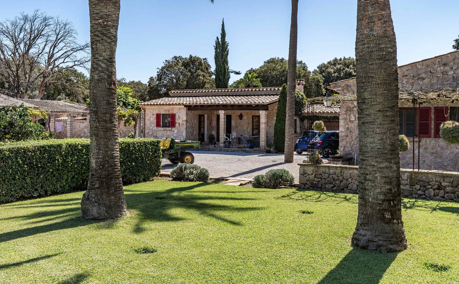 Villa rustique en pierre avec volets rouges et toit en tuiles, entourée d'une pelouse verdoyante et de palmiers.