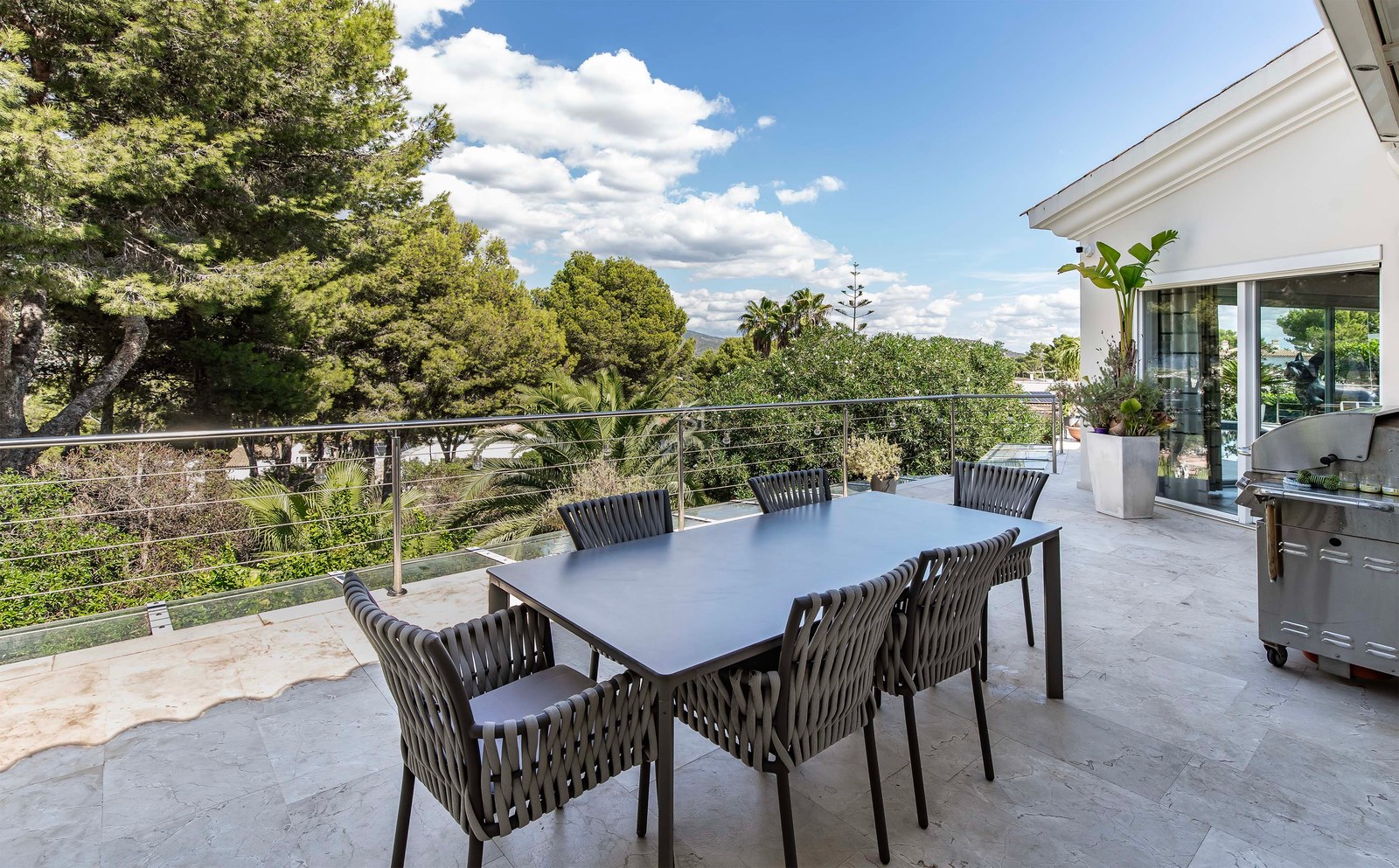 Terrasse extérieure avec salle à manger moderne, barbecue en inox et garde-corps en verre surplombant une forêt.